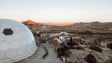 A desert landscape with a large geodesic dome structure, a dirt path, and a small building in the distance.