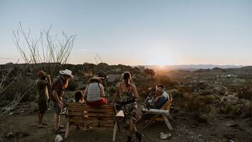 Un grupo de personas sentadas en bancos en un paisaje desértico con el sol poniéndose al fondo.