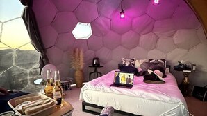 Una camera da letto con soffitto esagonale, un letto con un portatile, un comodino con bevande e vista dalla finestra.