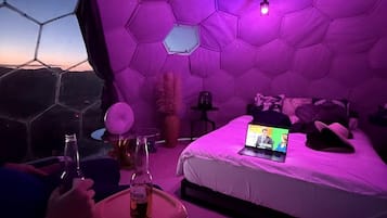 A room with a hexagonal ceiling, a bed with a laptop, and a bottle on a table.