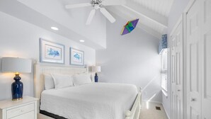 Ein Schlafzimmer mit einem weißen Bett, zwei Nachttischen mit Lampen, einem Deckenventilator und gerahmten Kunstwerken an der Wand.