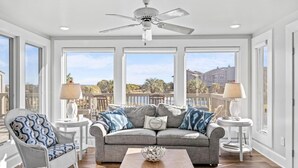 A spacious living room with a grey sofa, a rocking chair, and a coffee table. There are two side tables with lamps. The room has large windows offering a view of the outside.