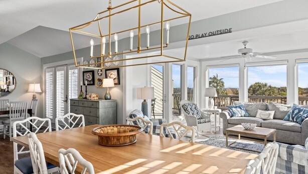 A spacious living room with a wooden dining table, white wicker chairs, a grey sofa, and a ceiling fan.