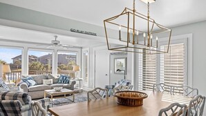 A spacious living room with a wooden dining table, comfortable seating, and a large window with blinds.