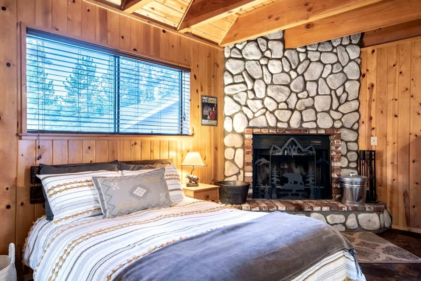 Una habitación acogedora con chimenea de piedra, paredes de madera, una cama con ropa de cama a rayas y un ventanal con persianas.