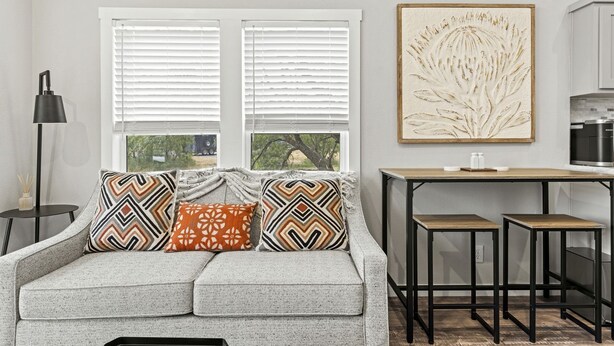 Una sala moderna con un sofá gris, cojines estampados, una mesita auxiliar, una lámpara de pie y una zona de cocina con mostrador y armarios.