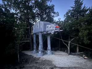 A large, metallic structure resembling a vehicle, elevated on a platform with support legs, surrounded by trees.