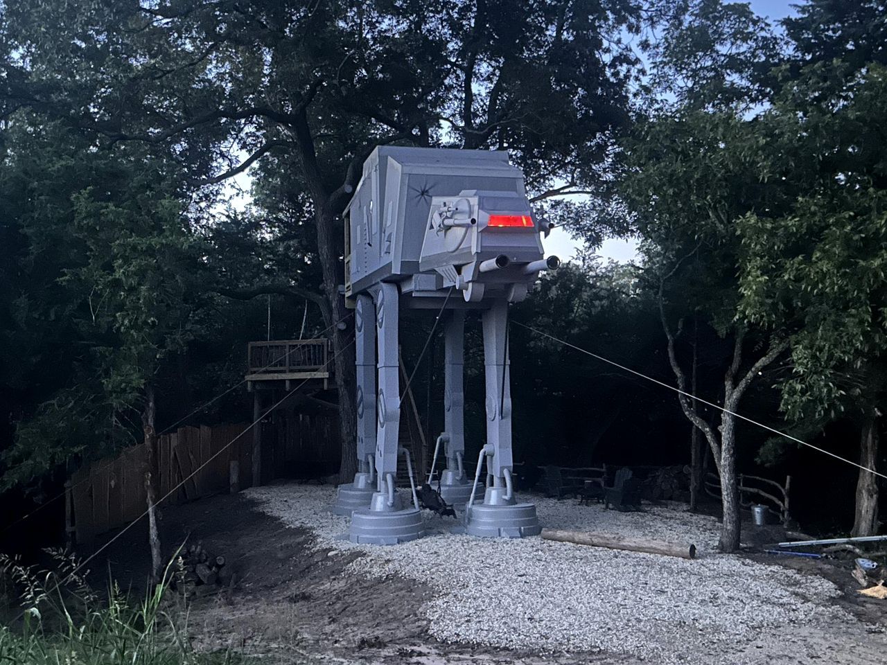 Une grande structure mécanique grise, ressemblant à un véhicule et soutenue par des pieds en métal, se trouve dans un espace extérieur boisé.