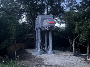 A large, gray, mechanical structure resembling a vehicle, supported by metal legs, in a wooded outdoor area.