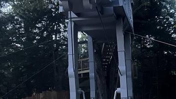 A large, grey, mechanical structure resembling a robot or vehicle, with a red light on top, set in a wooded area with a wooden fence and gravel ground.
