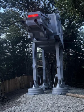 A large, grey, mechanical structure resembling a robot or vehicle, with a red light on top, set in a wooded area with a wooden fence and gravel ground.
