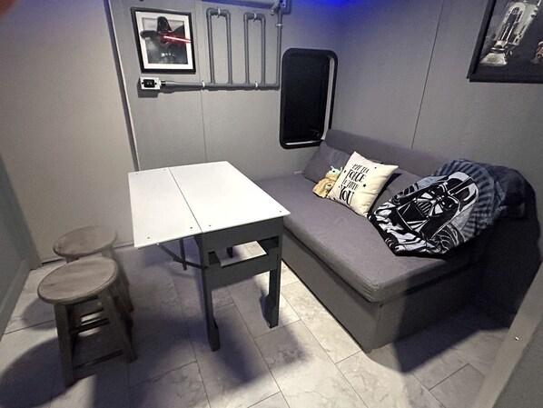 Une petite pièce avec un canapé gris, une table blanche et un petit tabouret. Il y a deux coussins sur le canapé, dont l’un est orné d’un motif de Star Wars.