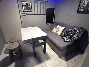Ett litet rum med en grå soffa, ett vitt bord och en liten pall. Det ligger två kuddar i soffan, varav en har ett Star Wars-motiv.