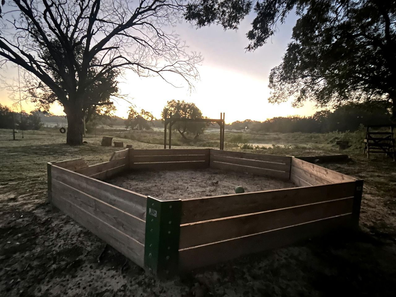A wooden raised garden bed in a rural setting with trees and a clear sky.