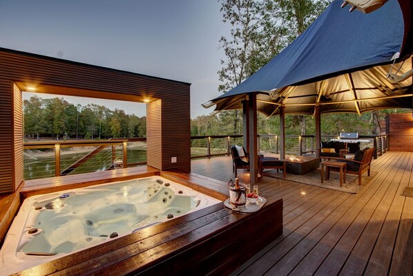 Un bain à remous sur une terrasse en bois, avec vue sur une rivière, des meubles d’extérieur et un espace barbecue.