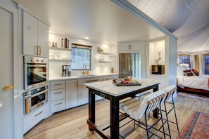 A modern kitchen with built-in appliances, a dining area with a table and bar stools, and a bedroom with a bed and curtains.