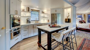Une cuisine moderne dotée d’appareils électroménagers intégrés, un coin repas avec une table et des tabourets de bar, et une chambre à coucher avec un lit et des rideaux.