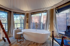 A spacious room with a large bathtub, wooden flooring, and large windows with curtains.