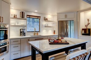 A modern kitchen with white cabinetry, a marble countertop, stainless steel appliances, and a window with a forest view.