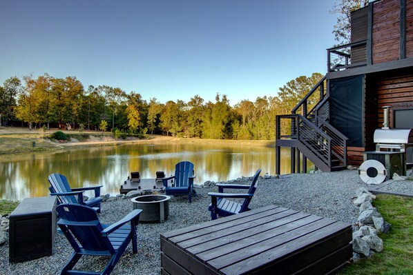 A lakeside deck with Adirondack chairs, a fire pit, and a grill.
