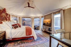 A bedroom with a large bed, a ceiling fan, a TV, a small table, and a chair.