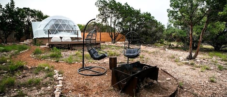 Un espace extérieur moderne aménagé avec deux chaises noires, un petit foyer et, au fond, une structure en forme de dôme transparent.