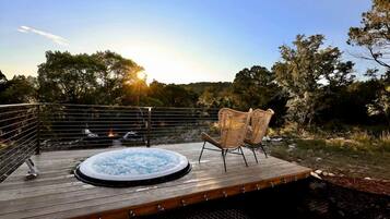 A wooden deck with a hot tub and wicker chairs, overlooking a natural landscape with trees and a body of water.