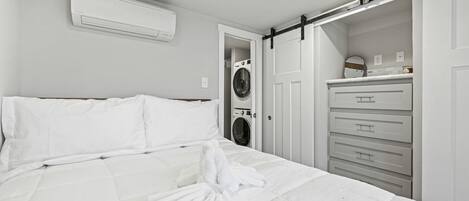 A hotel room with a bed, a white air conditioner, a washing machine, and a sliding closet door.