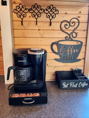 A coffee maker with a KEURIG logo, a coffee cup sign, and a box labeled 'But First Coffee'.