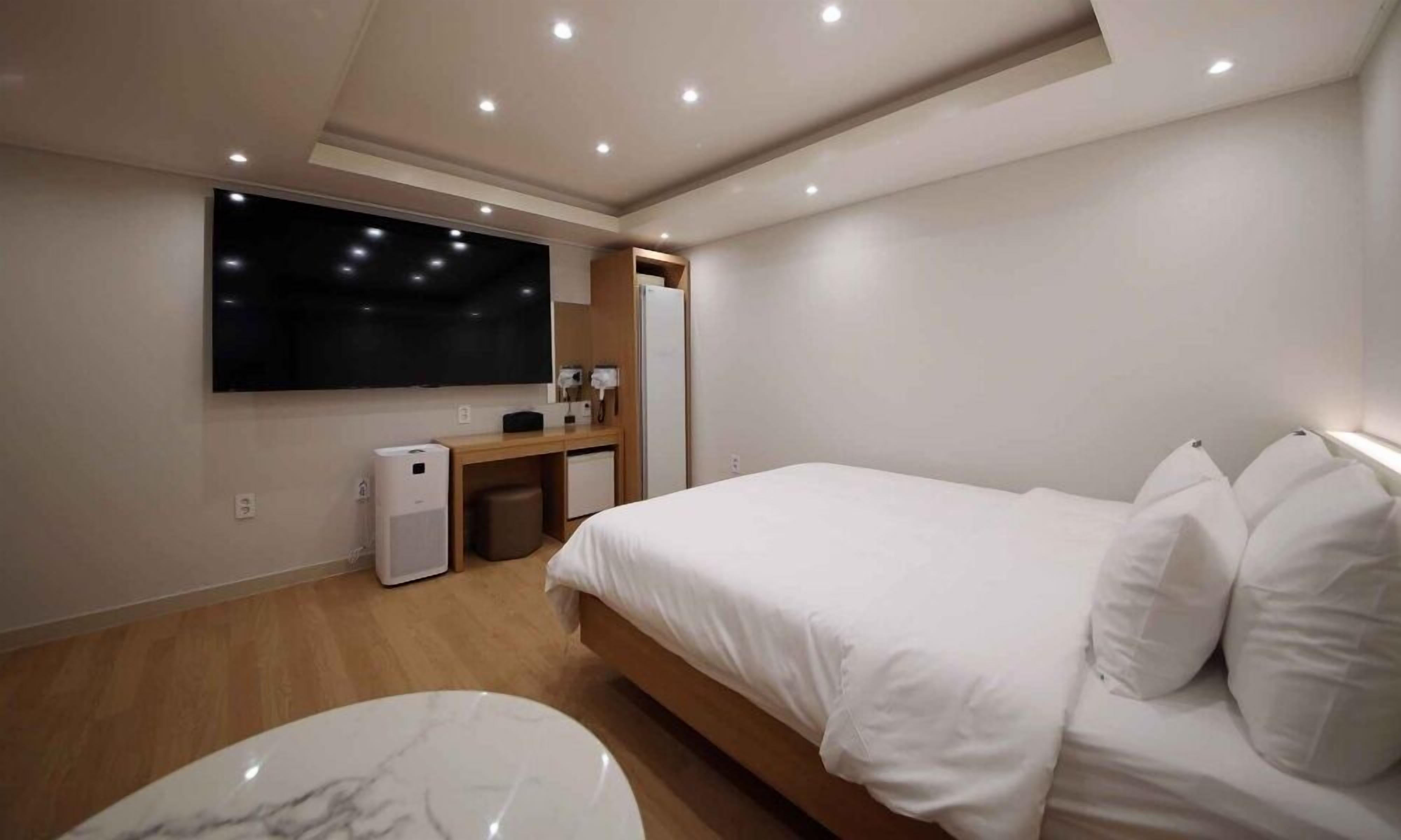 Una habitación de hotel moderna con una cama grande, un televisor de pantalla plana montado en la pared, un escritorio pequeño con una silla y un aire acondicionado compacto.