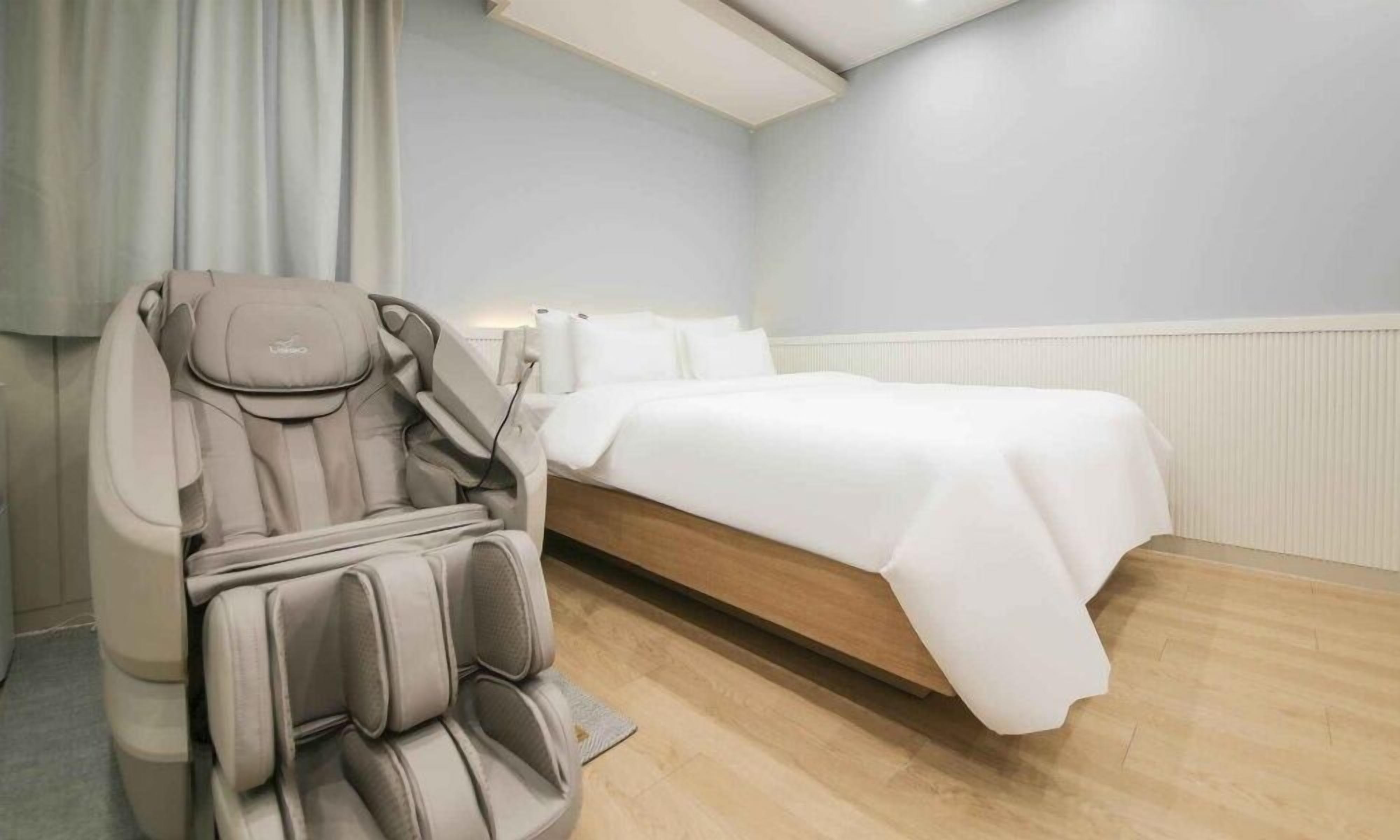 A room with a massage chair, a bed, and a wooden floor.