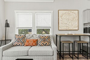 Un salon moderne avec un canapé gris, des coussins à motifs, une table d’appoint, un lampadaire et un coin cuisine doté d’un plan de travail et de placards.