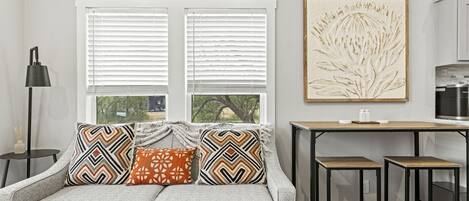 Una sala moderna con un sofá gris, cojines estampados, una mesita auxiliar, una lámpara de pie y una zona de cocina con mostrador y armarios.
