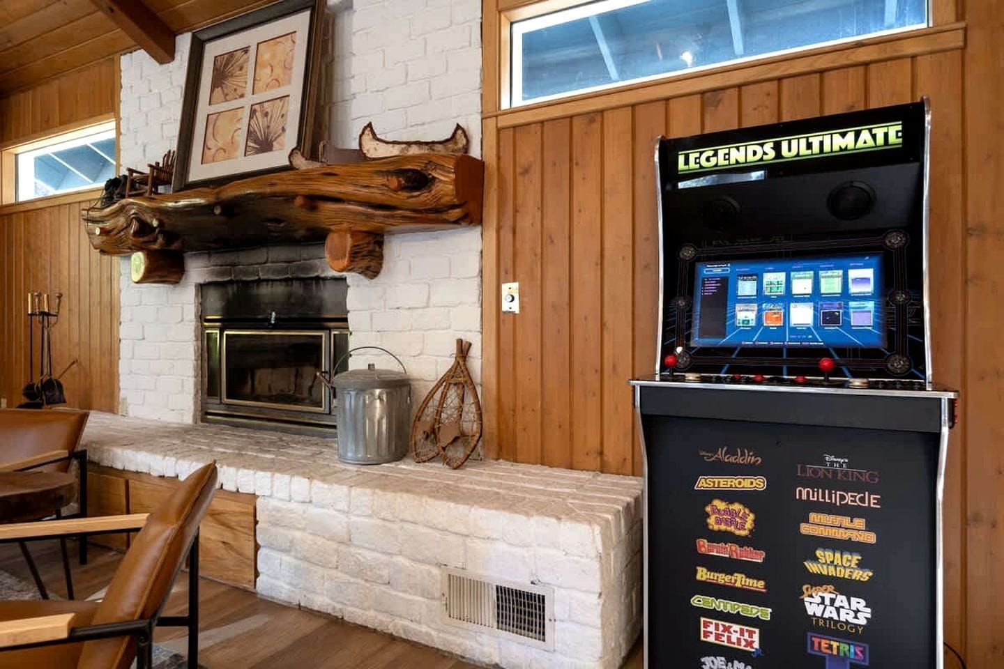 A wooden interior with a fireplace, a table with chairs, and a video game machine labeled 'LEGENDS ULTIMATE' with various game logos.