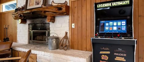 Un interior de madera con chimenea, una mesa con sillas y una máquina de videojuegos con la etiqueta 'LEGENDS ULTIMATE' y varios logotipos de juegos.