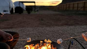 A campfire with marshmallows, a tent, and a wooden structure in the background.