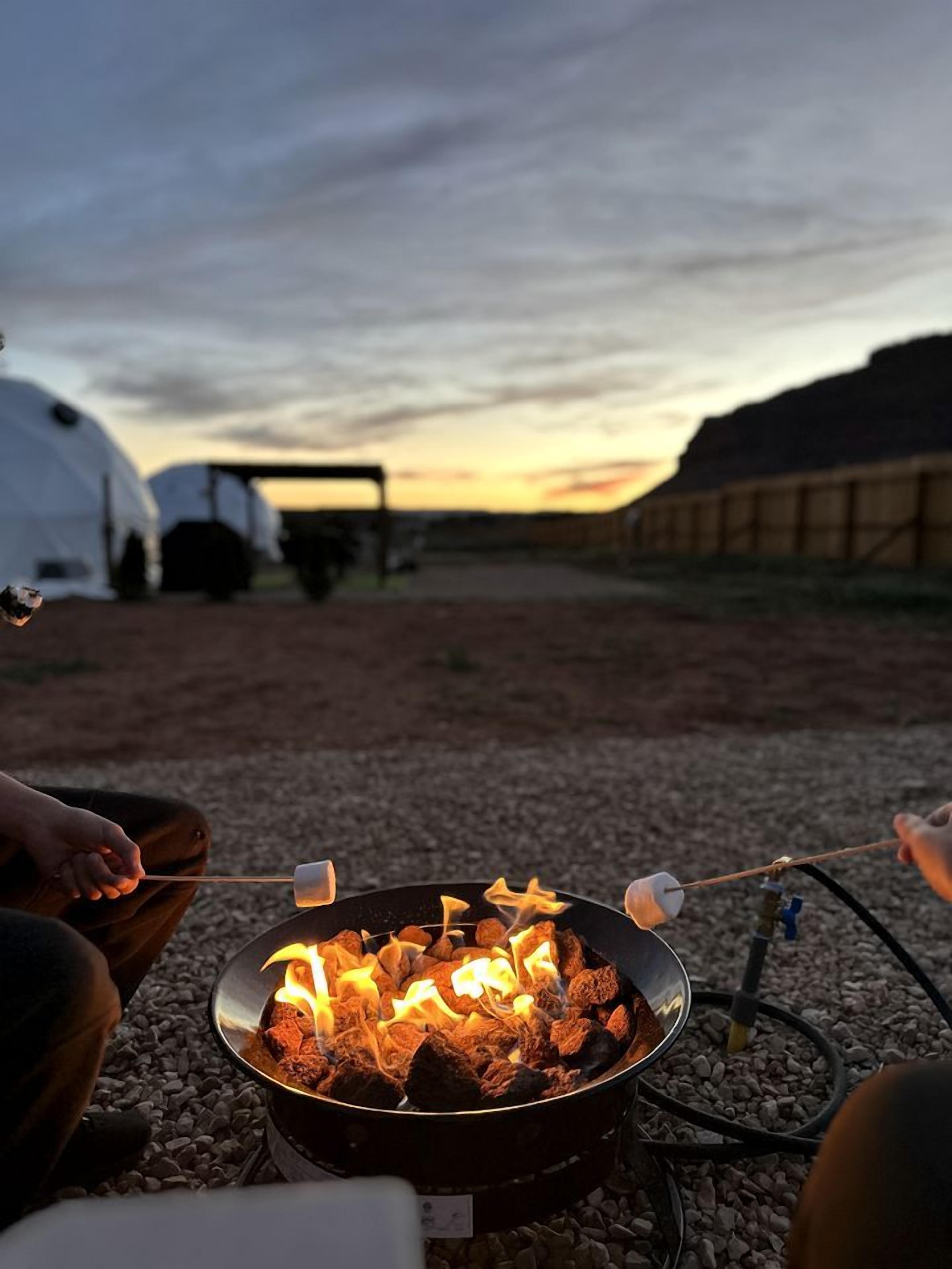 A campfire with marshmallows, a tent, and a wooden structure in the background.
