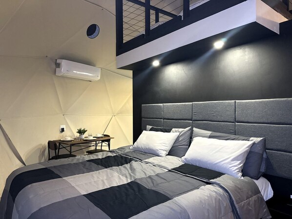 A modern bedroom with a large bed, a small table with a lamp, and a wall-mounted air conditioner.