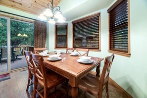 A dining area with a wooden table set for four, surrounded by chairs, with wooden blinds on the windows and a chandelier above. - Comfy Cabin with Outdoor Living Space with Spa in Big Bear Lake (Big Bear Lake)
