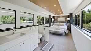 Une chambre moderne avec un grand lit, une salle de bain avec des toilettes et un lavabo, et de grandes fenêtres donnant sur un paysage verdoyant.