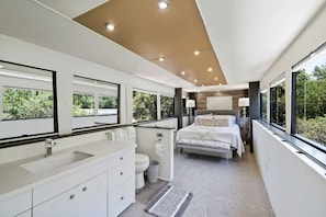 Un dormitorio moderno con una cama grande, un baño con inodoro y lavamanos, y ventanas amplias que ofrecen vistas al verdor.