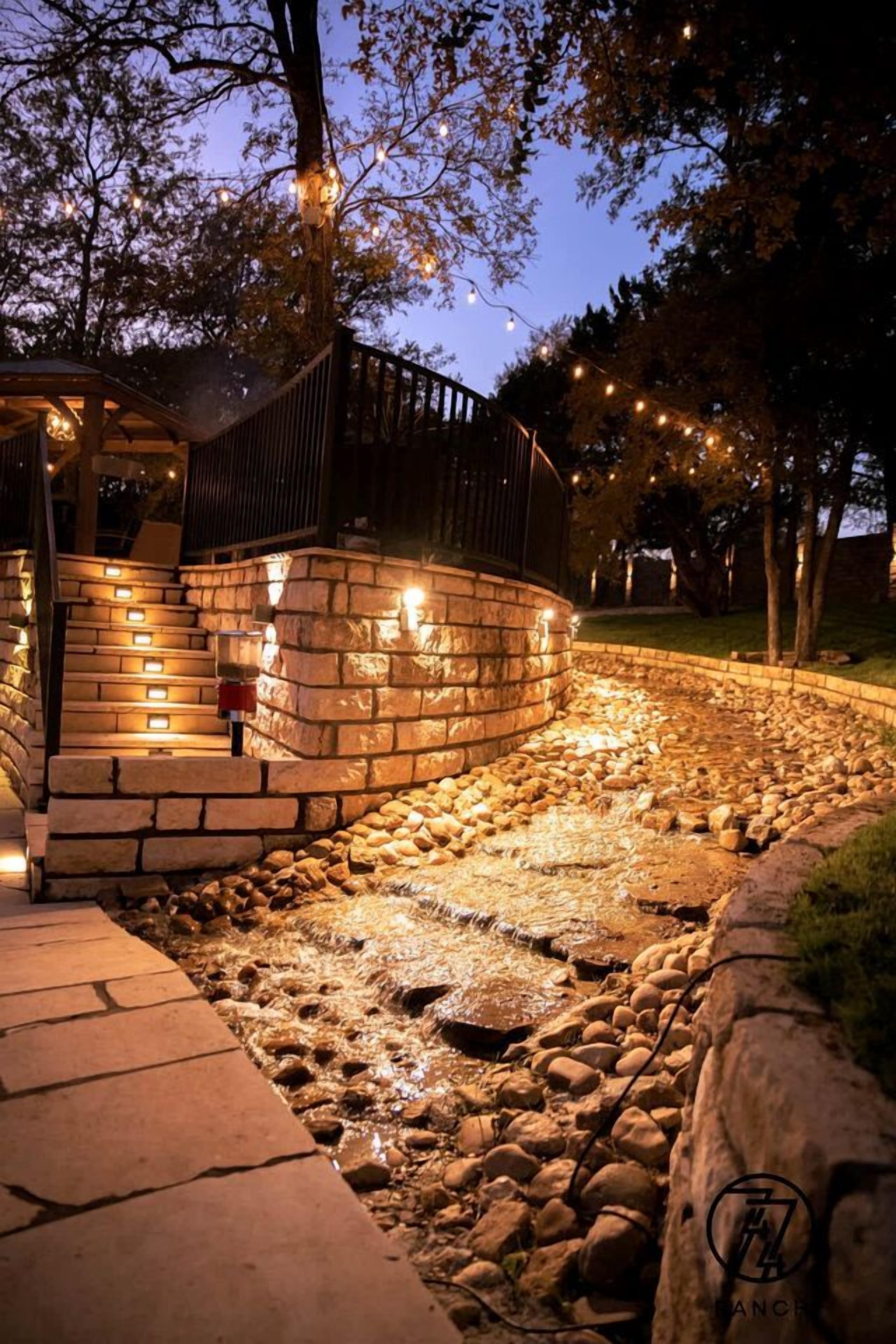 Een pad van stenen leidt naar een houten terras met lichtjes, omgeven door een aangelegde tuin.