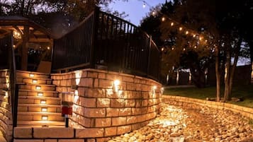 Een pad van stenen leidt naar een houten terras met lichtjes, omgeven door een aangelegde tuin.