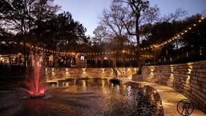 Un espace extérieur entouré de murs en pierre, agrémenté d’un élément décoratif aquatique, de guirlandes lumineuses et d’une petite construction.