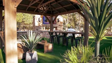 Een terras buiten met een houten pergola, groene stoelen en een eettafel.