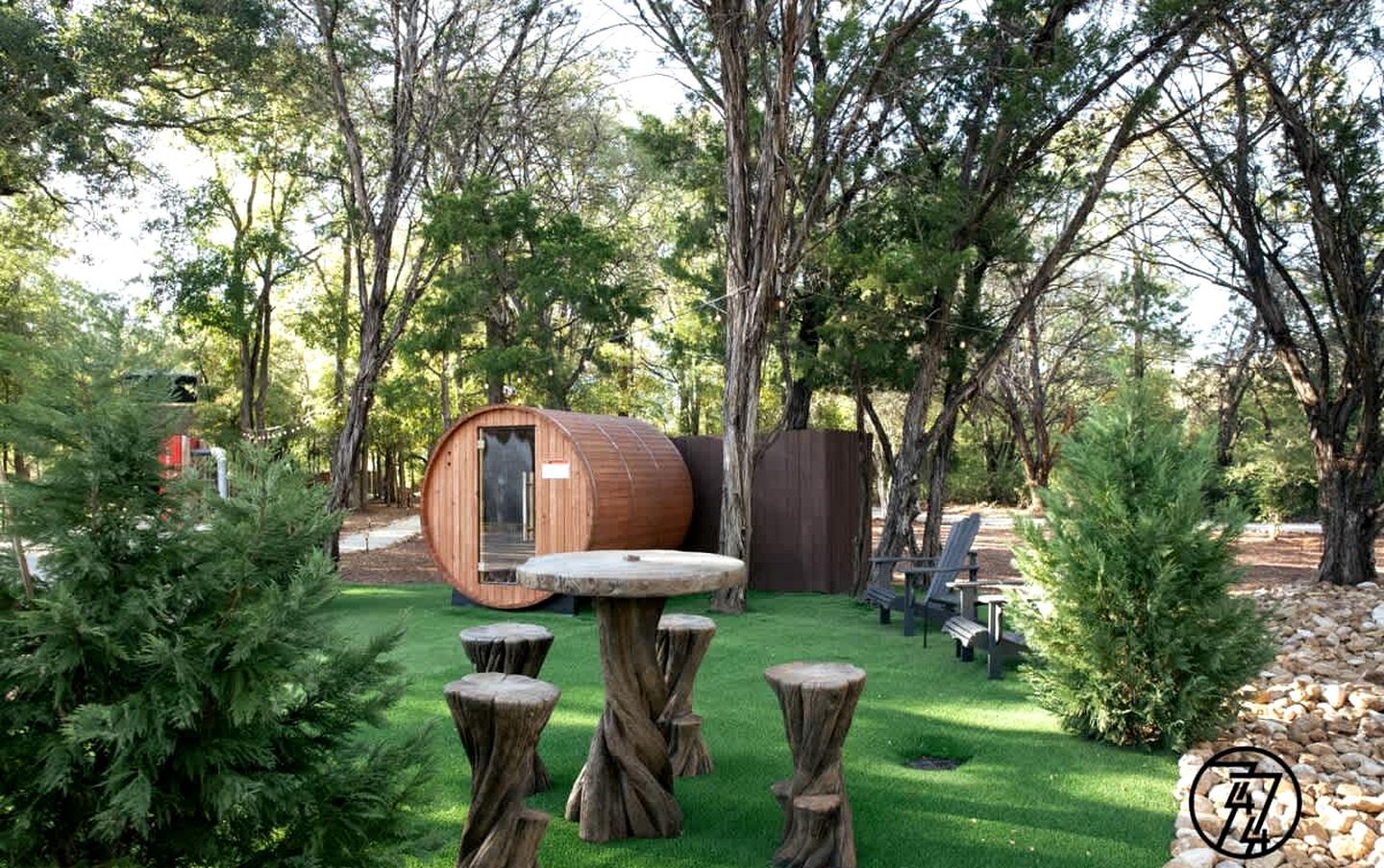 Un décor extérieur rustique composé d’une structure en forme de tonneau en bois, d’une table ronde et de plusieurs tabourets en bois.