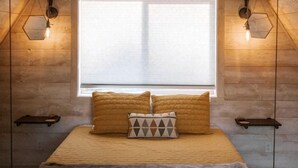 Ein Schlafzimmer mit schräg stehender Decke, einem großen Fenster, einem Bett mit vielen Kissen und Wandleuchten.