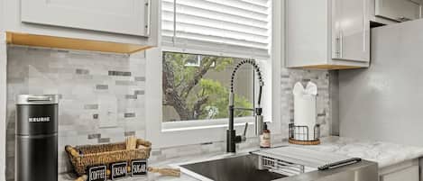Cocina moderna con un lavamanos de acero inoxidable, una cafetera Keurig y una cesta de mimbre con café, crema y azúcar.