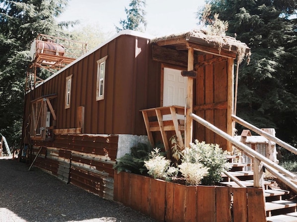 A rustic wooden building with a staircase leading to a door, surrounded by greenery.
