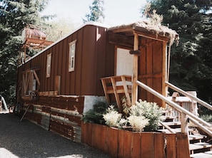 A rustic wooden building with a staircase leading to a door, surrounded by greenery.